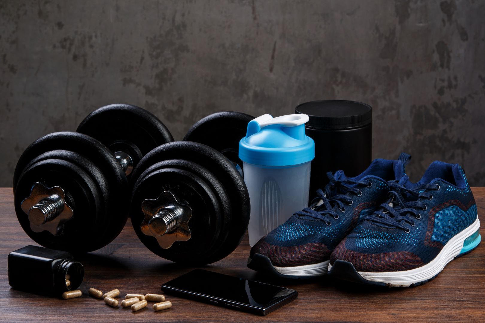 Dumbbells, running shoes, supplements, and a shaker bottle on a gym floor representing sports nutrition and performance support